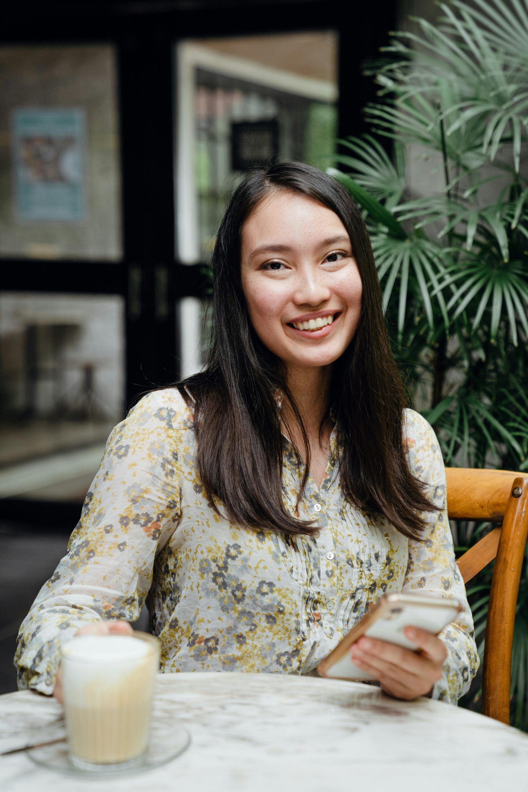 Cheerful young Asian lady with mobile phone in casual clothes smiling and drinking latte while chilling in outdoor cafe