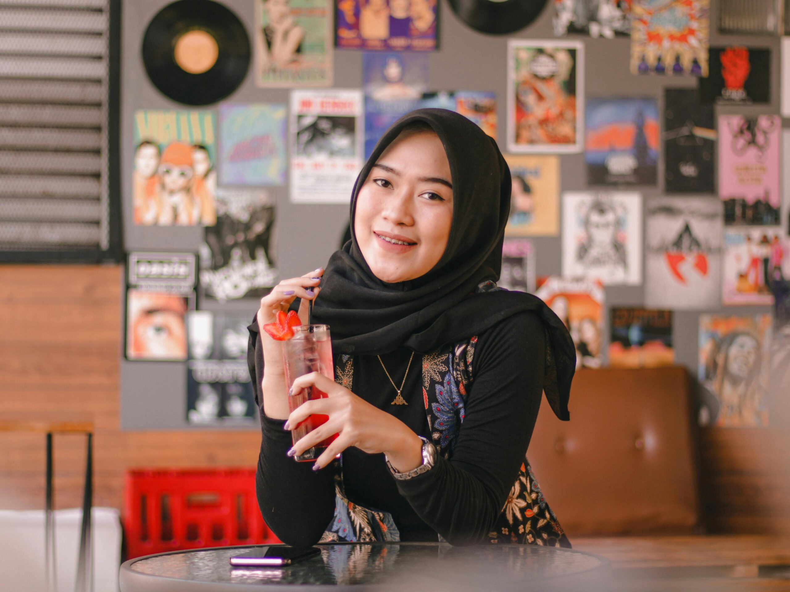 A smiling woman in a hijab enjoying a drink indoors with vibrant posters in the background.